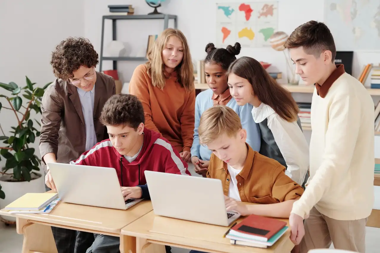 A group of diverse students collaborating on laptops in a classroom setting, guided by a teacher.
