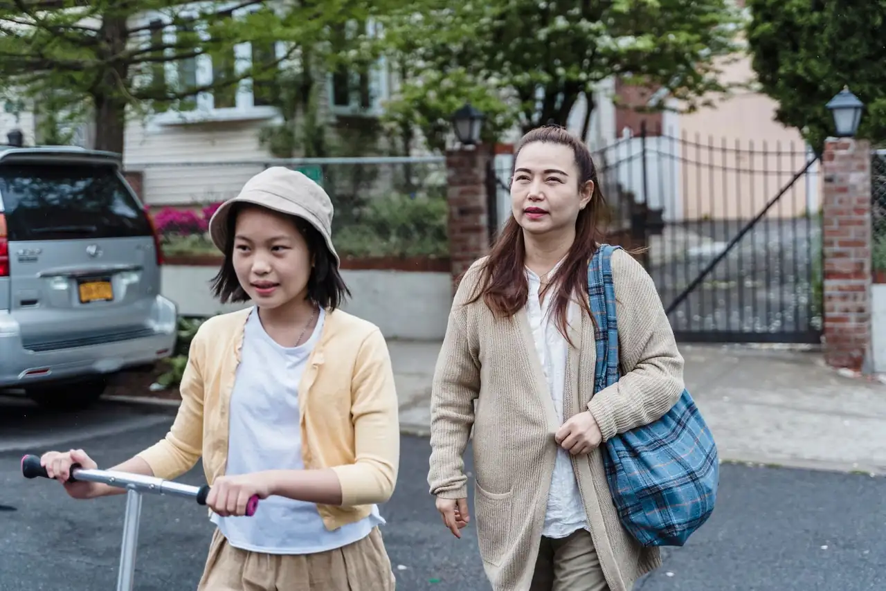 A mother and daughter enjoy a walk with a scooter in an urban neighborhood setting.